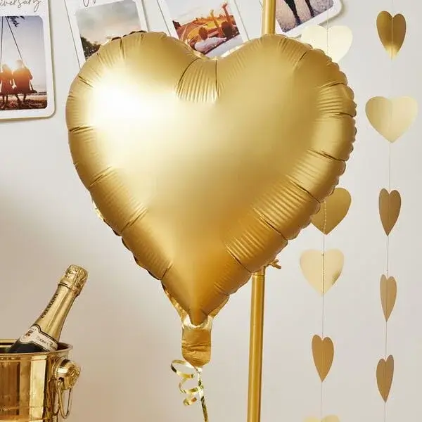 Gold heart-shaped foil balloon on a stand with a champagne bottle in an ice bucket and hanging heart decorations in the background.